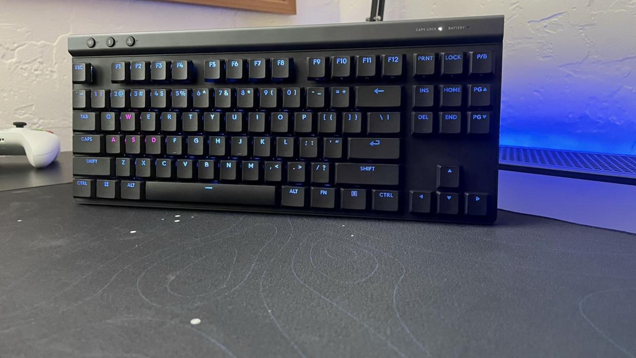  The Logitech G515 LIGHTSPEED TKL gaming keyboard standing on a black desk next to a game controller. 