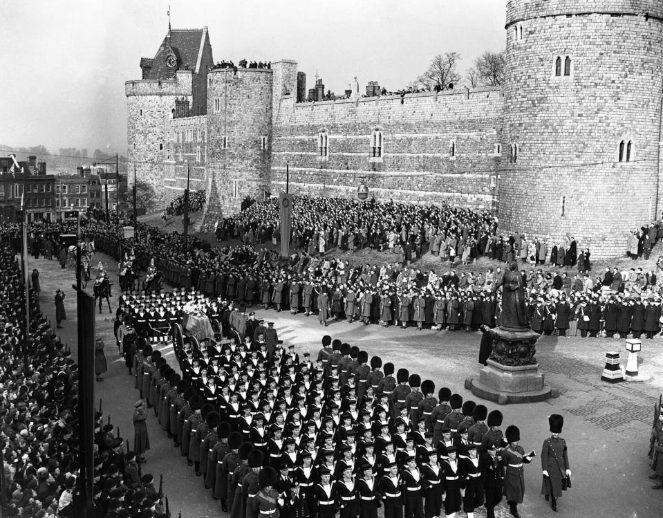 Men in uniform march in formation near crowds lined up and clustered in groups outside a castle