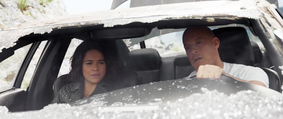 Two actors in an action scene inside a damaged car, appearing focused