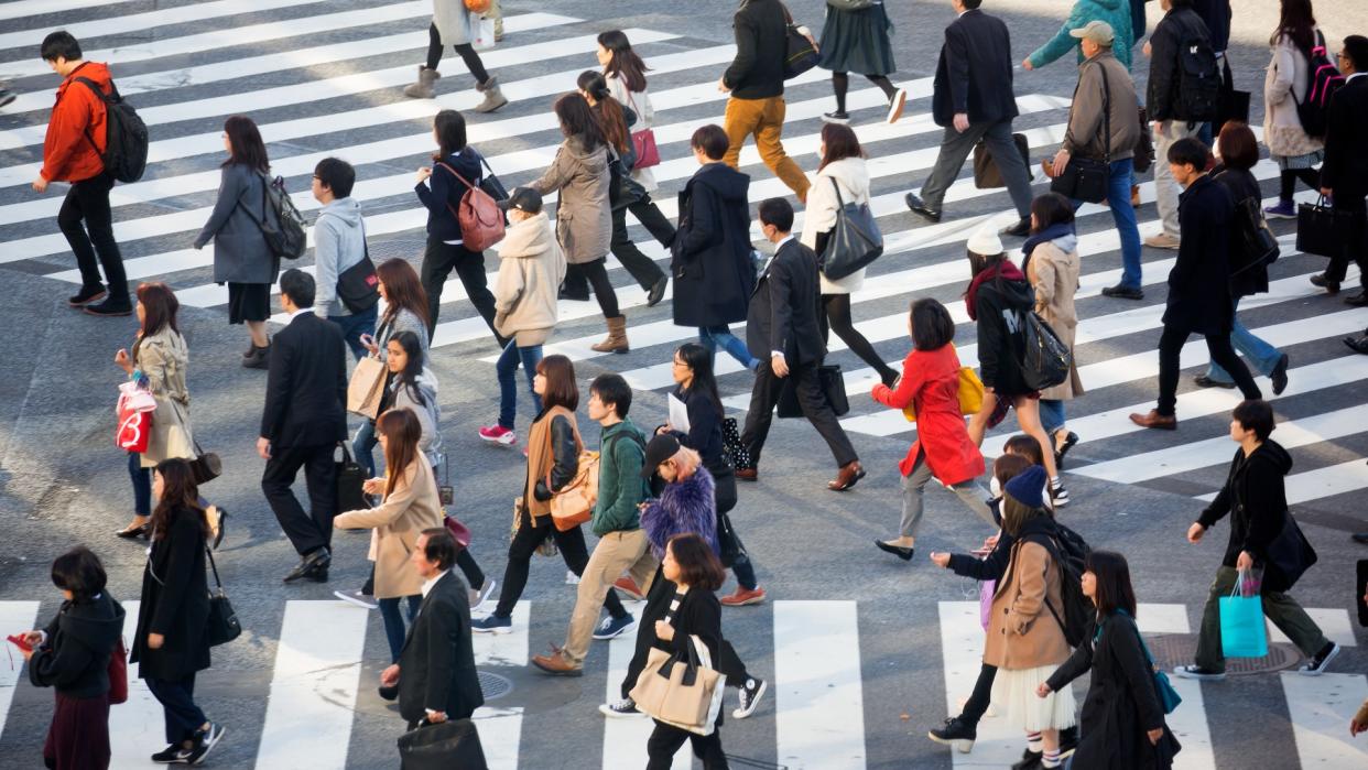  Image of a crowd of people crossing the Shibuya intersection. One set of white marking lines can be seen running parallel to the bottom side of the image, while another run diagonally towards the upper left-hand corner of the image. A range of people can be seen crossing from those carrying backpacks, others carrying shopping bags, some dressed in suits and others in jeans. 