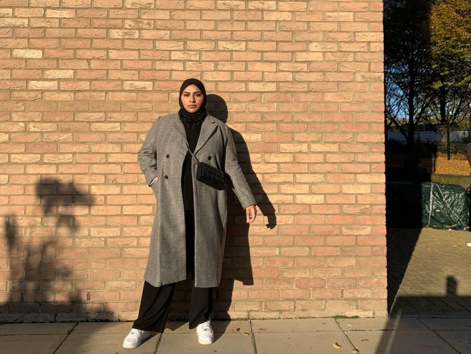 Faiza Zeria stands, looking serious against a tan brick wall with one hand in her coat pocket. She wears a black hijab, grey wool coat, long black pants, and white sneakers.