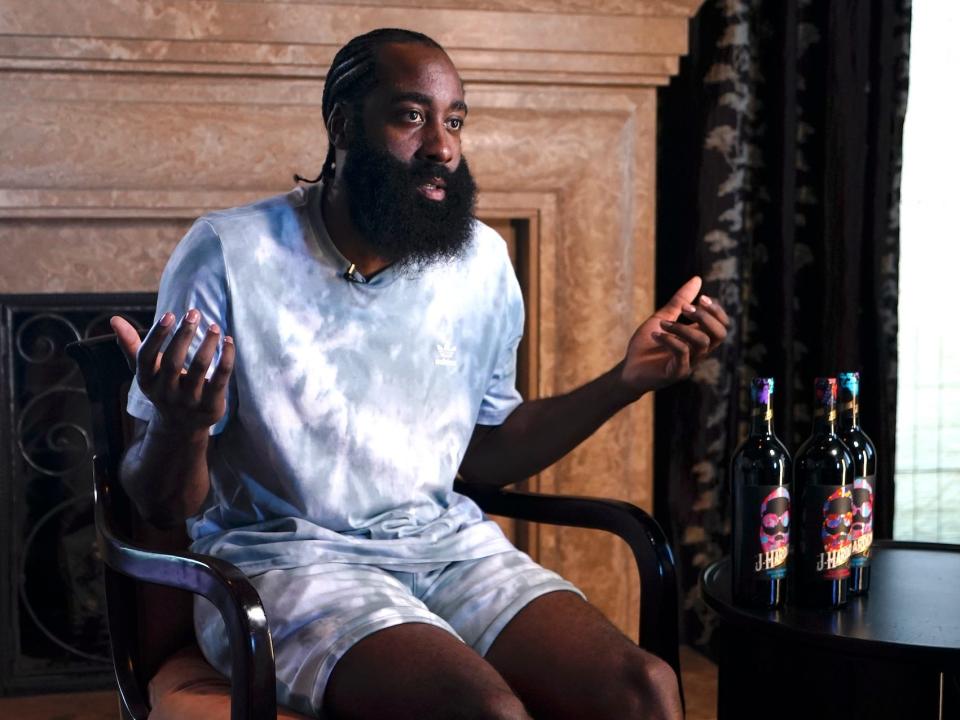 James Harden raises his hands while sitting next to 3 bottles of his wine.