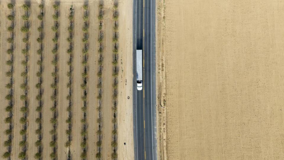 Aerial view of a tractor trailer on a paved road driving between farmland plots.