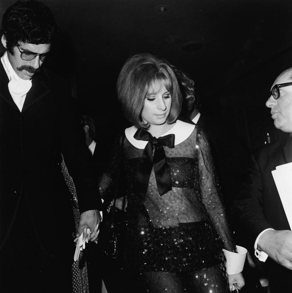 Barbra in a sequined black jumpsuit with a white collar and bow and pockets placed over her chest. The rest of the outfit is see-through.