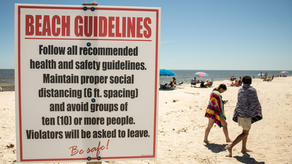 A guidelines sign at a public beach includes "maintain proper social distance" and "avoid groups of 10 or more people"