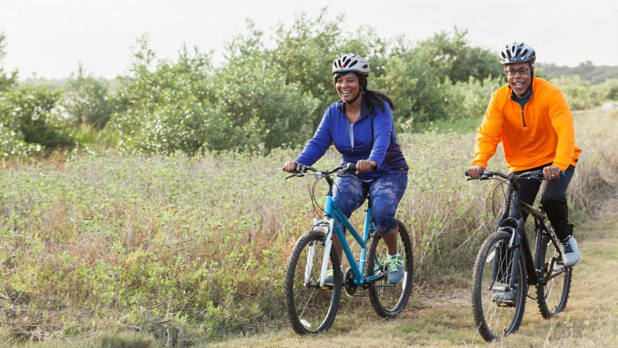  photo shows a man and woman riding bikes next to each other on a grassy path. They're both wearing helmets and smiling 