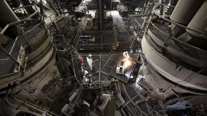 Workers in white suits and helmets inside a vast power plant, seen from above.