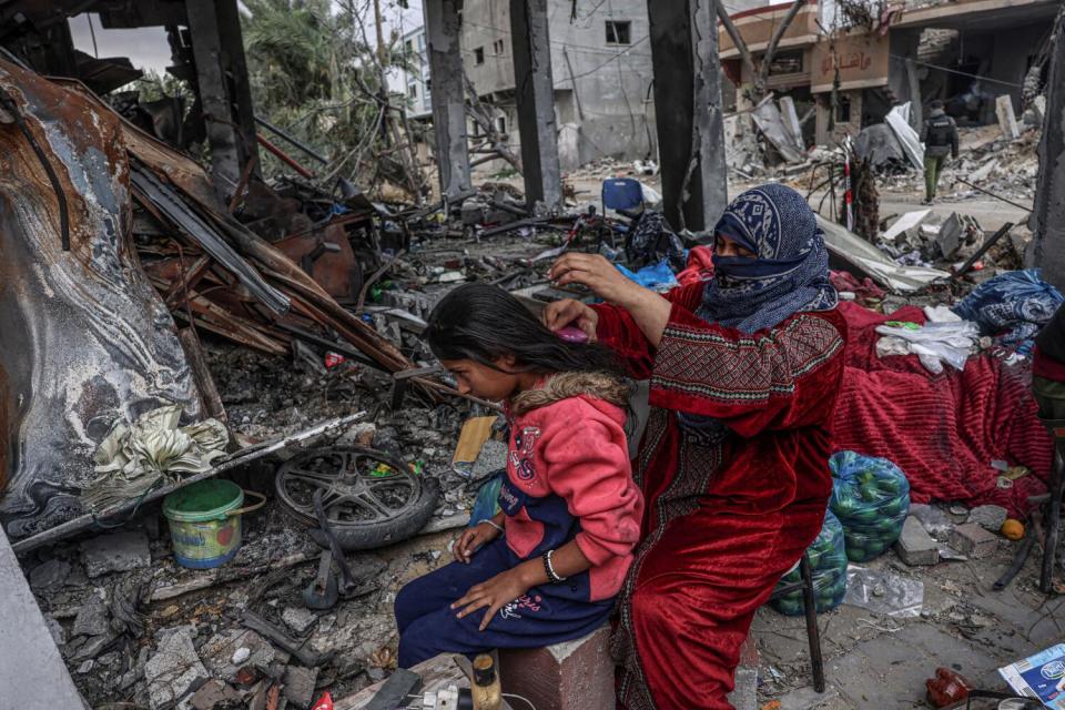 A woman in a red robe, her face covered, brushes the hair of a girl in a pink top, also seated, amid a scene of devastation