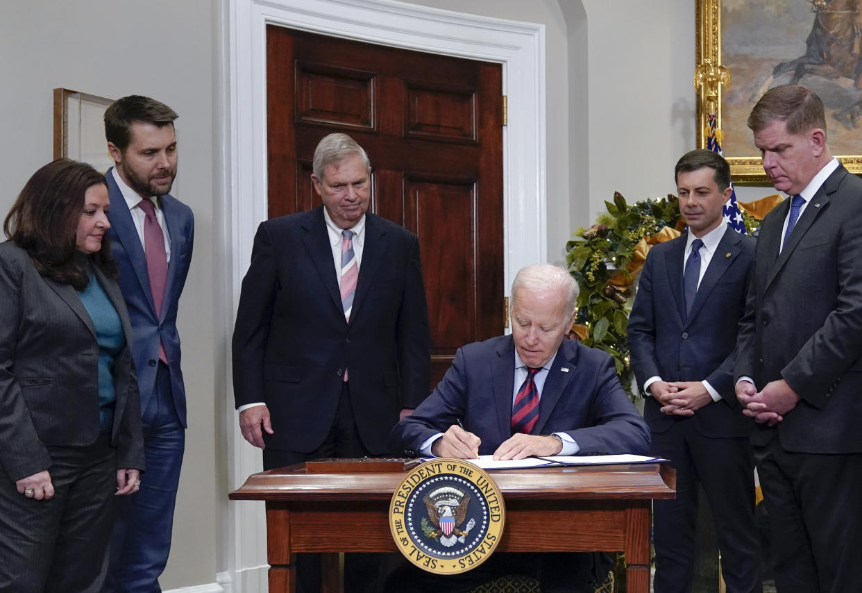 President Biden signs the rail strike bill at a desk, flanked by Director of Made in America at the Office of Management and Budget Celeste Drake, National Economic Council Director Brian Deese, Agriculture Secretary Tom Vilsack, Transportation Secretary Pete Buttigieg, and Labor Secretary Marty Walsh. 