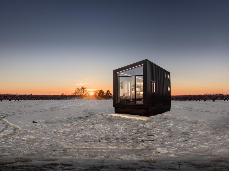 a black tiny home with a large glass wall on a snowy field at sunset