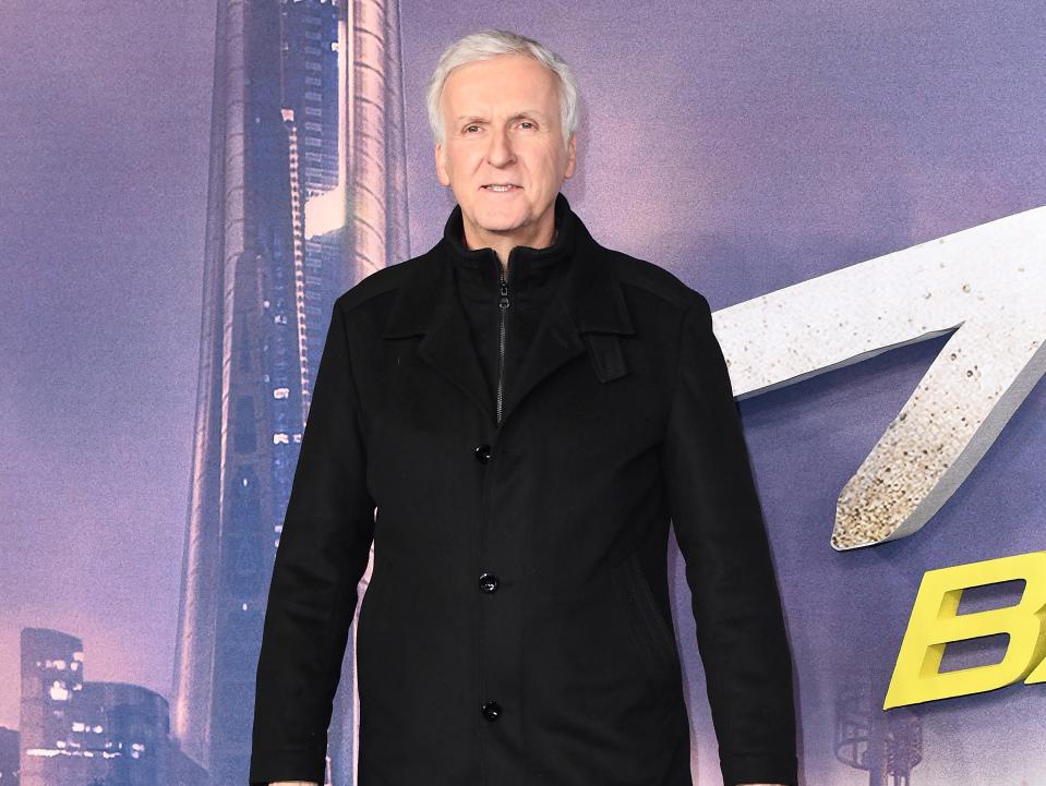 james cameron attending the world premiere of alita: battle angel in london wearing a black jacket, zip-up underneath, and stanidng in front of a purple cityscape background