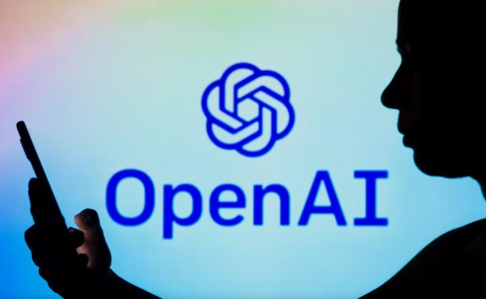 A photo illustration shows the OpenAI logo in the background of a silhouetted woman holding a cellphone. ChatGPT is a chatbot launched by developer OpenAI.