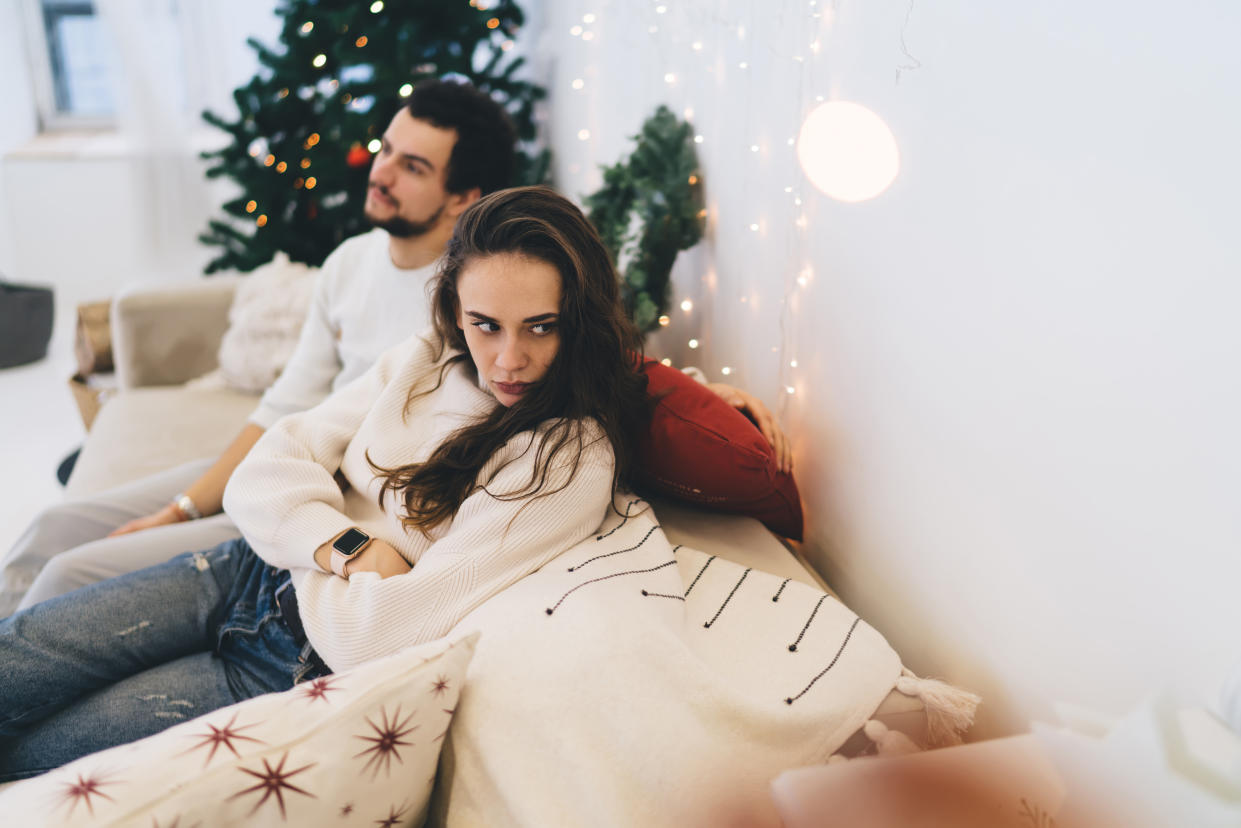 A woman crosses her arms and looks away while sitting on the same sofa as a man, who is sat next to her