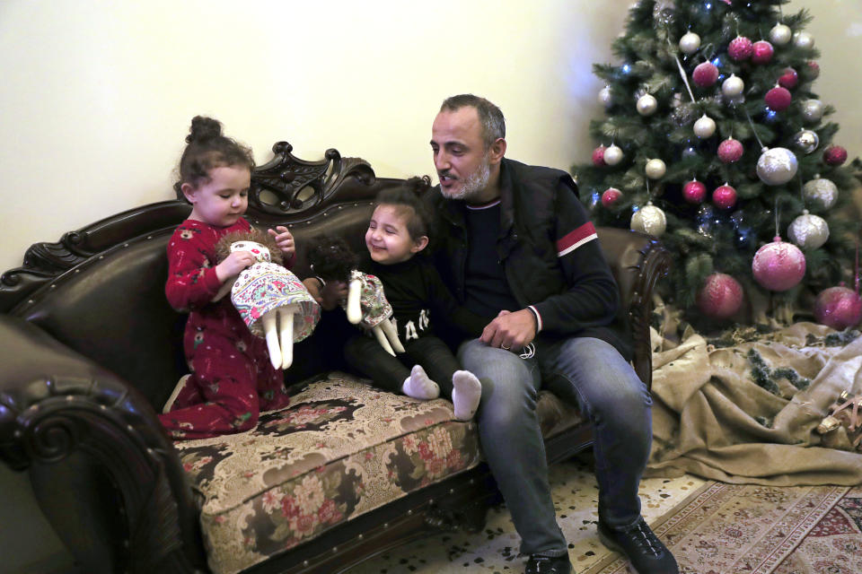 Georges Chlawuit, right, sits with his daughters Sama, 2, left, and Sima-Rita, 3, center, as they play with dolls at a relative's home, in Beirut, Lebanon, Tuesday, Dec. 29, 2020. In August, the massive explosion in Beirut blew out the windows of their family home. Painter Yolande Labaki made 100 dolls for children affected by the destruction. (AP Photo/Hussein Malla)