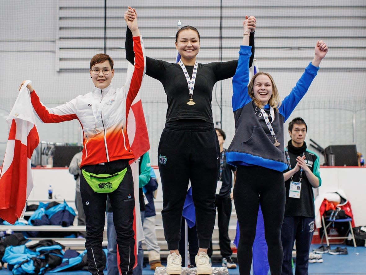 Danica Taylor of Team Northwest Territories, middle, stands atop the podium next to silver medallist Petra Amossen of Team Greenland, left, and bronze medallist Emma Noskey of Team Alberta North after winning gold in the one-foot high kick on Day 4 of the 2023 Arctic Winter Games in Fort McMurray, Alta., on Wednesday. (Evan Mitsui/CBC - image credit)