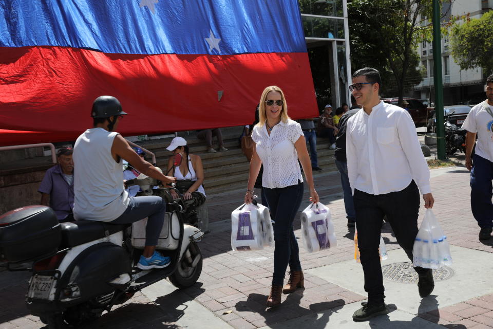 A couple, carrying their purchases of toilet paper and bottled water, walk through a plaza where opponents of the Nicolas Maduro government are gathering for an opposition rally in Caracas, Venezuela, Saturday, May 11, 2019. Opposition leader Juan Guaidó has called for nationwide marches protesting the Maduro government, demanding new elections and the release of jailed opposition lawmakers. (AP Photo/Rodrigo Abd)