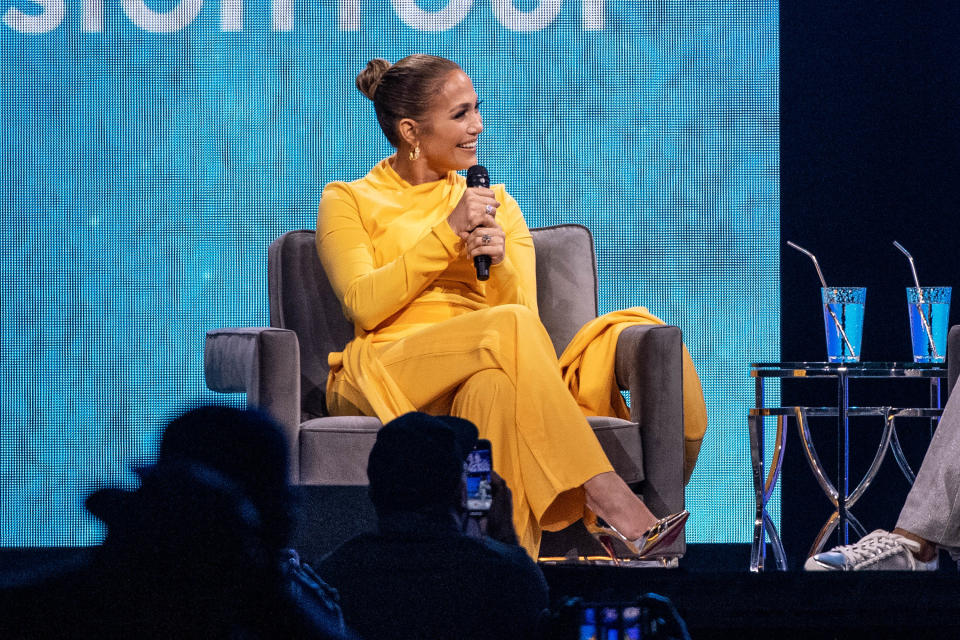 Jennifer Lopez is wearing a yellow suit, sitting in an armchair on stage and holding a microphone