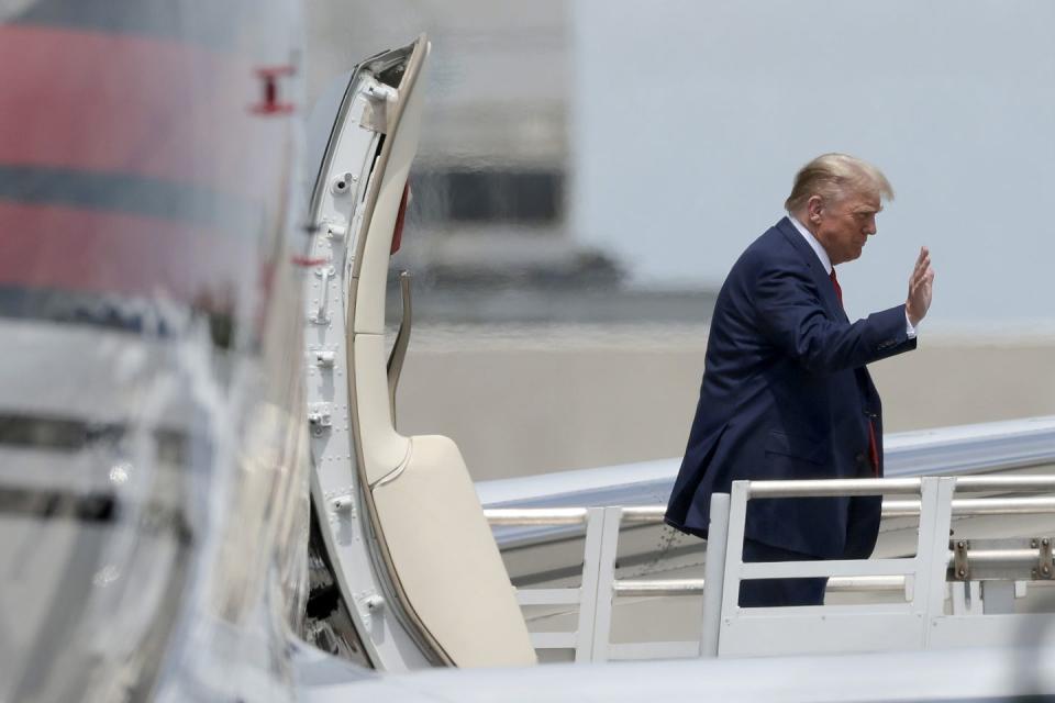 donald trump, wearing a blue suit, walks along the ramp of an airplane and waves off camera