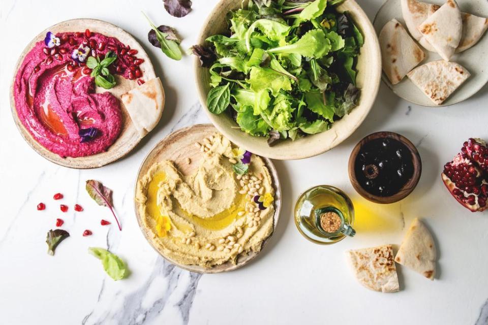 variety of homemade traditional and beetroot spread hummus with pine nuts, olive oil, pomegranate served on ceramic plates with pita bread and green salad on white marble background flat lay, space