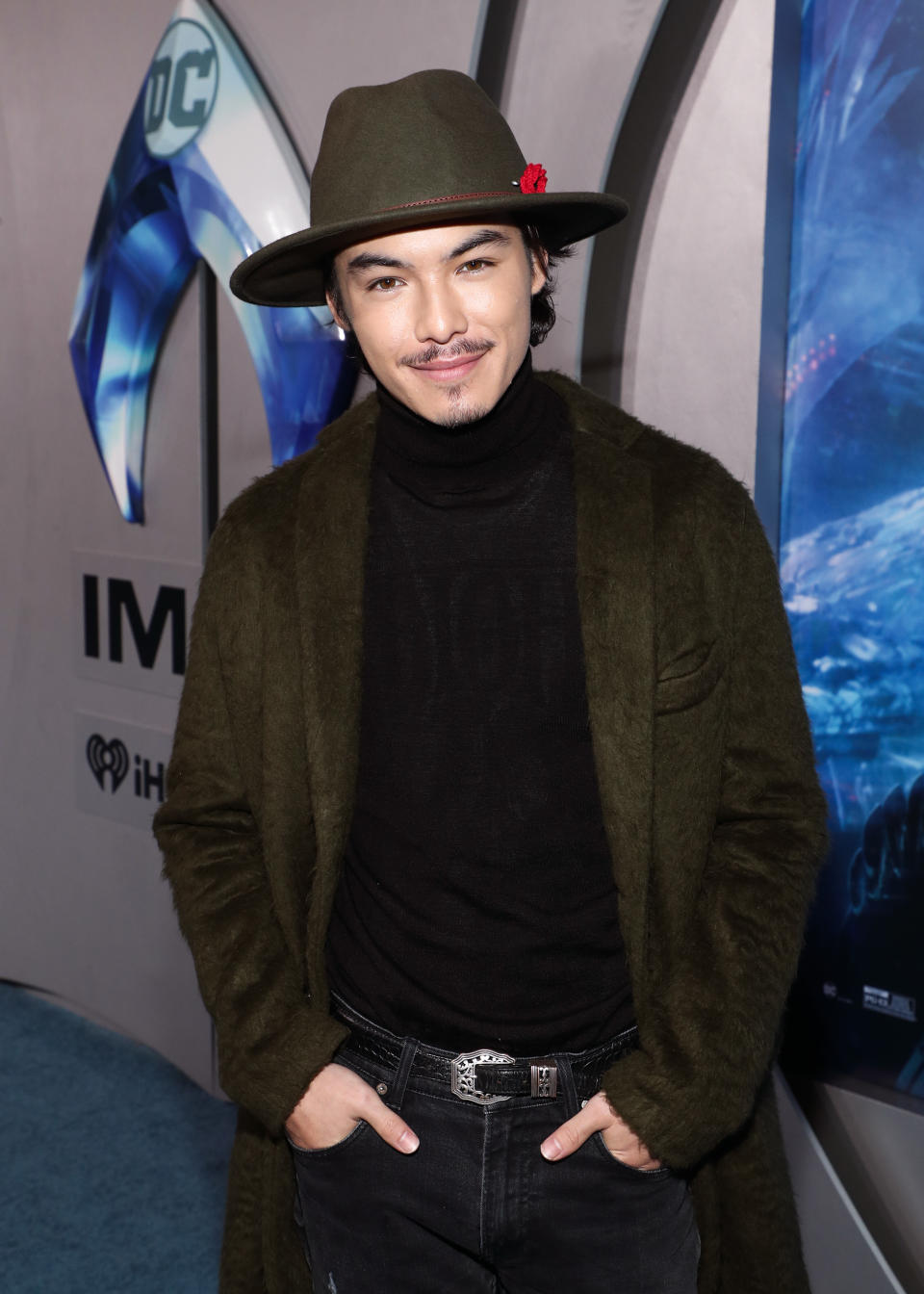 Ryan Potter in a dark turtleneck, green coat, and stylish hat smiles on the red carpet at a movie premiere