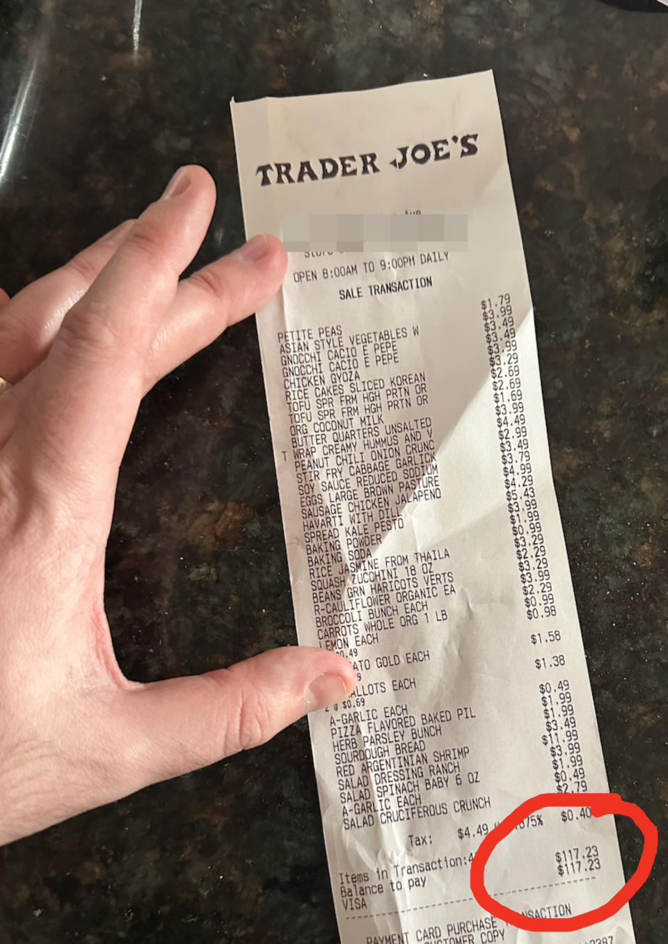 Hand holding a Trader Joe's receipt listing various grocery items and their prices, with a total of $117.43 at the bottom