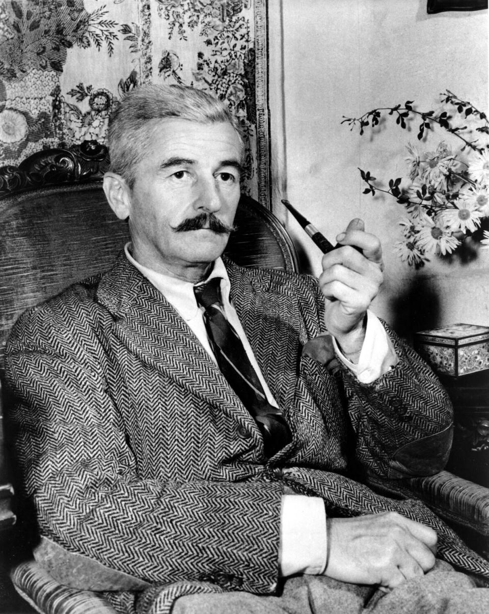 William Faulkner sits in a chair with a pipe in hand