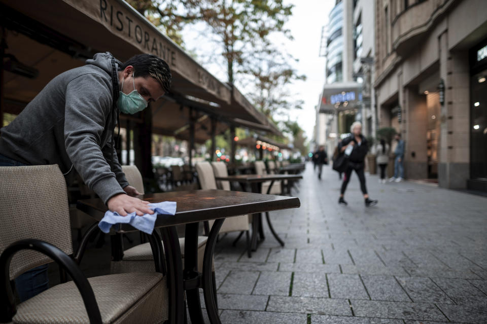 An employee wearing a mouth and nose protector wipes tables in a restaurant on the shopping street Koenigsallee in Duesseldorf, Germany, Friday, Oct. 9, 2020. The corona pandemic is increasingly becoming a problem in the large cities and conurbations of North Rhine-Westphalia. (Fabian Strauch/dpa via AP)