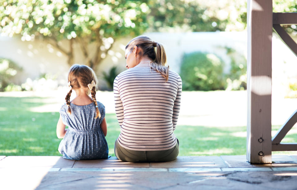 An adult woman and a young girl with braided hair sit on an outdoor porch step, facing away, enjoying a sunny garden view