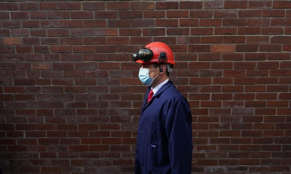 Paul Williams on a visit to an industrial site, wearing a hard hat, goggles and mask, photographed in profile against a brick wall