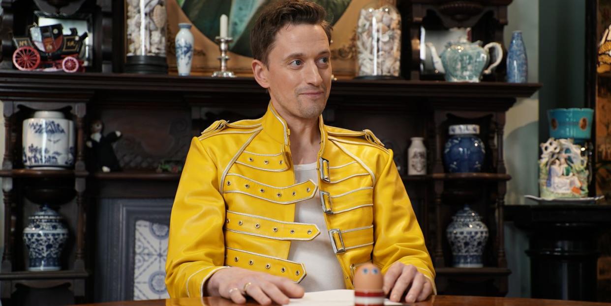 john robins sits inside the taskmaster house's task room, in front of a table containing a task and an egg in a striped cup with googly eye stickers on it