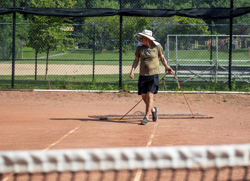 John Cornack a volunteer with Frick Park Clay Courts Tennis Club (FPCCTC), revitalizes the red clay tennis courts in Frick Park in the Pittsburgh neighborhood of Regent Square, on Monday, June 17, 2024. Cornack is a retired brain injury specialist from Michigan who takes care of the community tennis courts through the overwhelming heat by taking precautions with proper head gear. (Esteban Marenco/Pittsburgh Post-Gazette via AP)