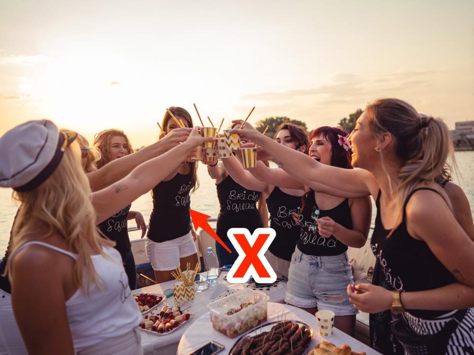 A group of women toast drinks on a boat during a bachelorette trip. A red x points to their shirts.