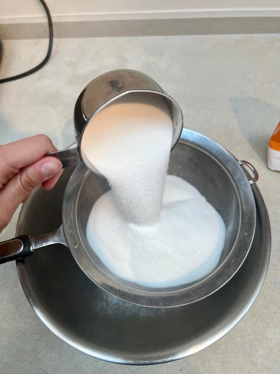 A cup of flour, sugar, and baking soda being poured into a sieve.