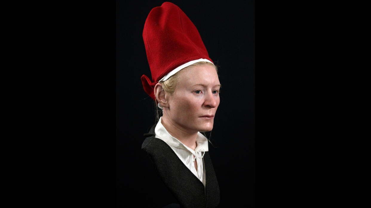  We see the reconstruction of a woman's face and upper torso. She has pale skin, blonde hair and a solemn expression. She wears a tall bright red hat, a gray jacket and a white blouse with a collar.  