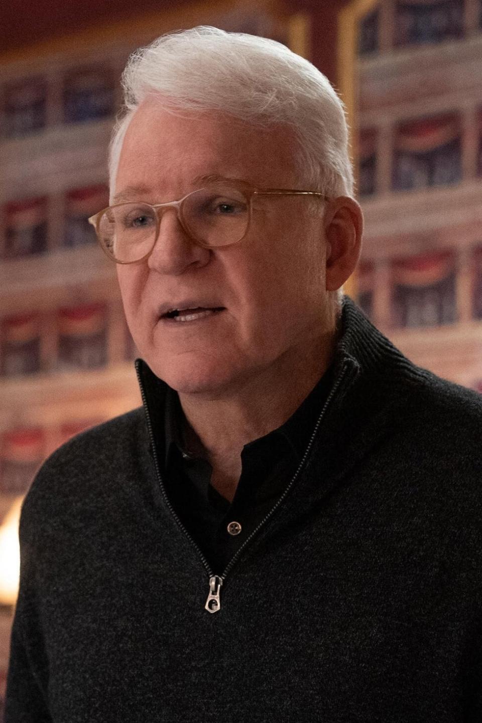 Steve Martin, wearing a dark zip-up sweater, is speaking and gesturing with his hands in an indoor setting decorated with intricate wallpaper