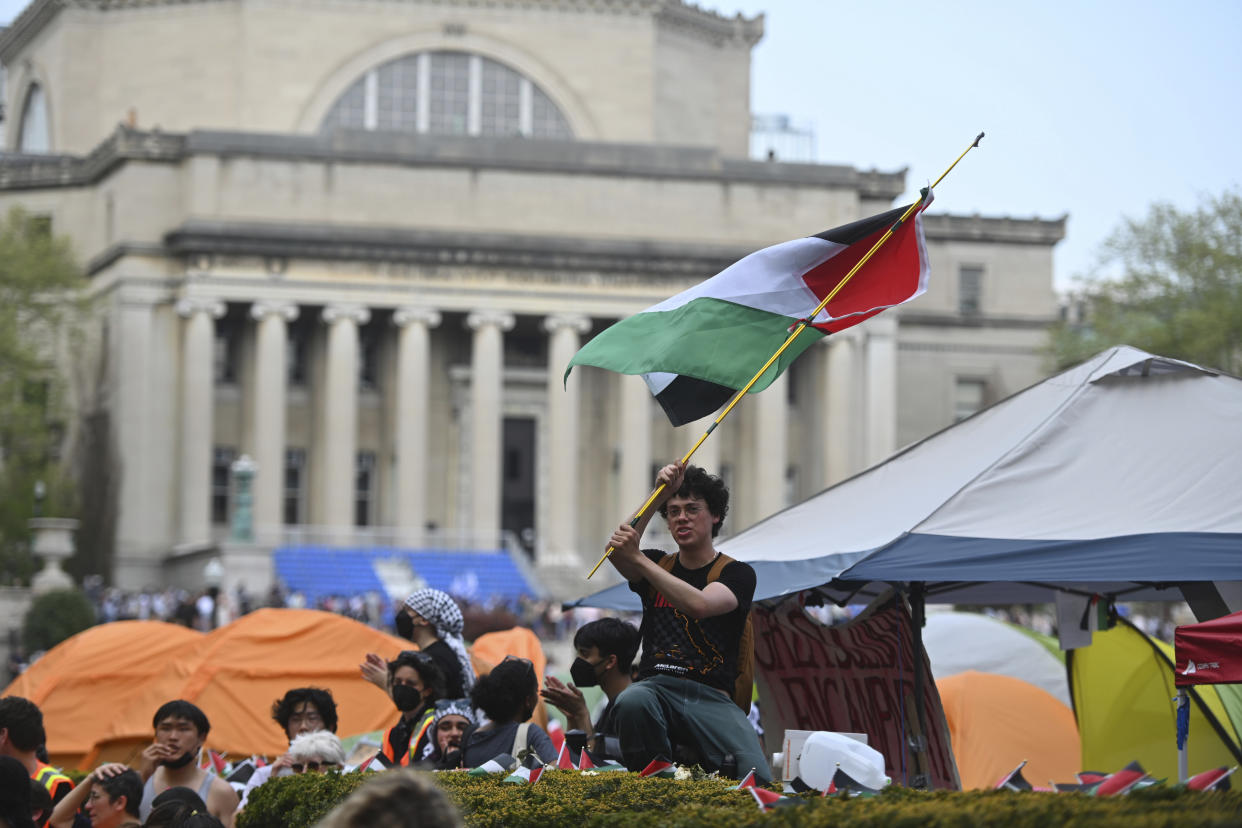 Several people are seen near tents in front of what appears to be a university building. One holds a Palestinian flag.