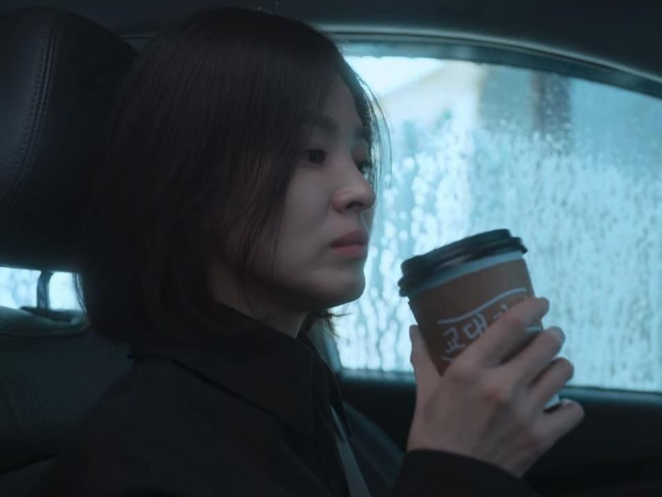 dong-eun in the glory holding a coffee cup in her right hand while going through a car wash. the cup has a brown sleeve on it, with the words gyodae cafe written on it in hangul