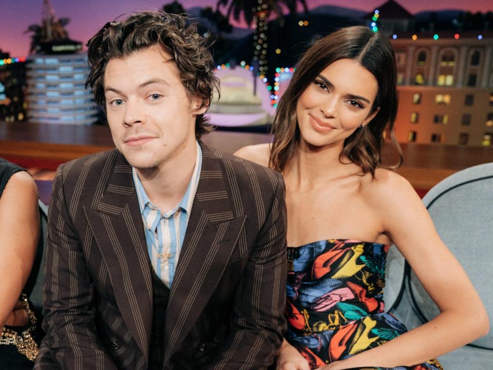 Harry smiling in a brown striped suit and Kendall sitting next to him smiling in a colorful strapless top.