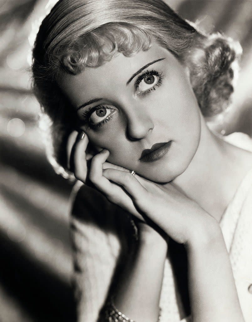 Bette Davis in a vintage black and white portrait, gazing thoughtfully with her chin resting on her hands. She has short, wavy hair and is wearing a light-colored top