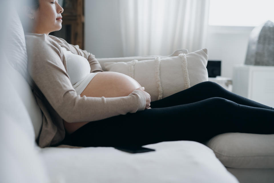 Asian pregnant woman with her eyes closed touching her belly, relaxing on sofa at cozy home. Expecting a new life, wellbeing, healthy pregnancy lifestyle