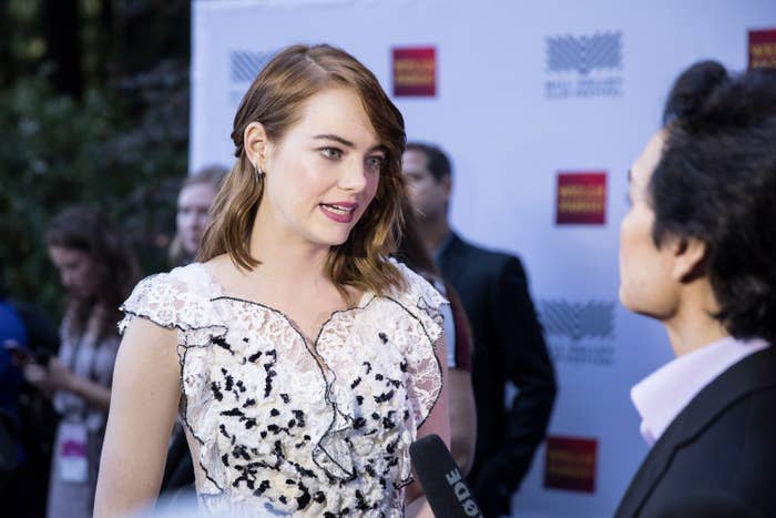 Emma Stone, in a lace dress with black floral details, talks to an interviewer at an event, with onlookers in the background