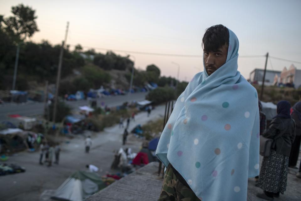 A migrant with a blanket stands on a rooftop of a building as others gather on a road leading from Moria to the capital of Mytilene, on the northeastern island of Lesbos, Greece, Thursday, Sept. 17, 2020. Greek police are moving hundreds of migrants to an army-built camp on the island of Lesbos Thursday after a fire destroyed an overcrowded facility, leaving them homeless for days. (AP Photo/Petros Giannakouris)