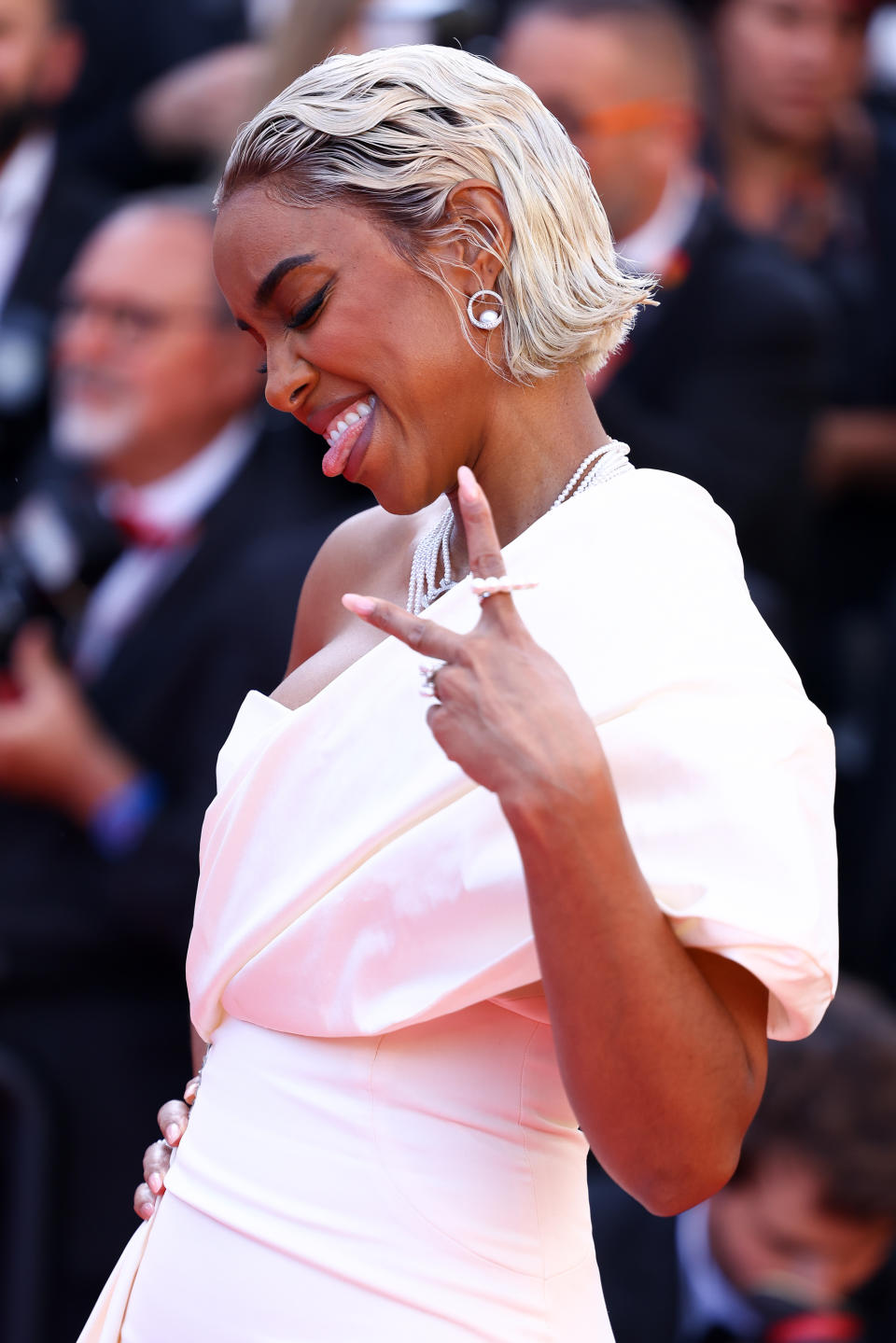 Actress Kelly Rowland poses playfully on the red carpet, making a peace sign and sticking her tongue out, dressed in a draped, elegant white gown