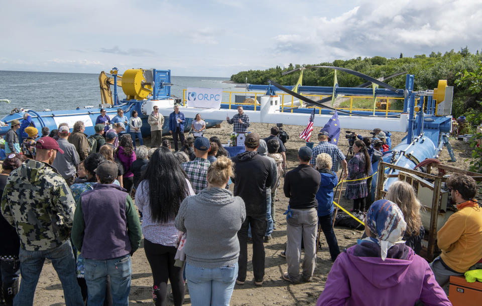 In this Tuesday, July 16, 2019, photo released by the Alaska Governor's office, Gov. Mike Dunleavy, center rear, speaks in front of a Riv-Gen Power System turbine on the bank of the Kvichak River in Igiugig, Alaska. A tiny Alaska Native village is adopting an emerging technology to transform the power of a local river into a renewable energy source. (Austin McDaniel/Alaska Governor's Office via AP)