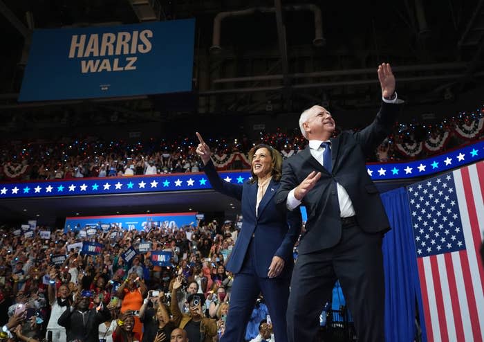 Kamala Harris and Tim Walz on stage at a rally, waving to a large crowd in an indoor venue with a "Harris Walz" banner above