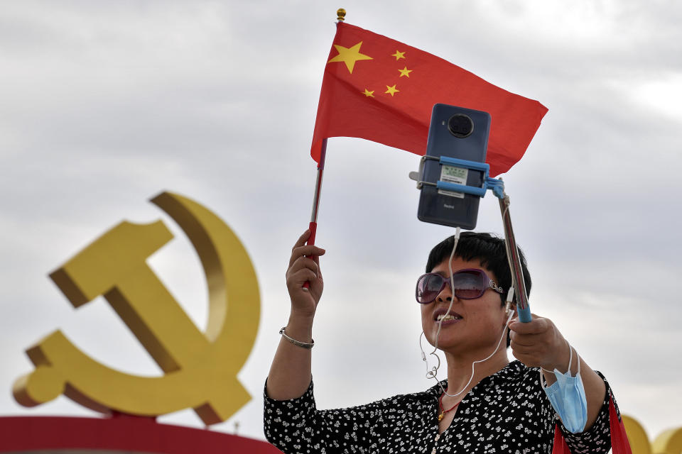 A woman holds a national flag while using a smartphone to film herself with a communist party's logo on display at Tiananmen Square to mark the 100th anniversary of the founding of the ruling Chinese Communist Party in Beijing on Monday, July 5, 2021. Chinese leader Xi Jinping on Tuesday attacked calls from some in the U.S. and its allies to limit their dependency on Chinese suppliers and block the sharing of technologies. (AP Photo/Andy Wong)