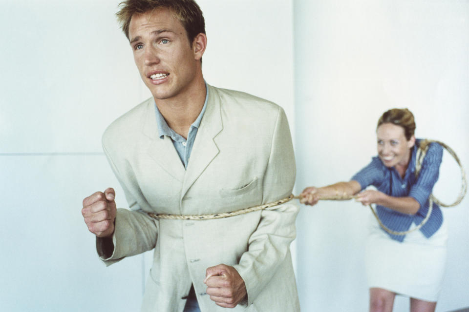 A man in a light suit is tied with a rope pulled from behind by a woman in office attire, creating a humorous struggle scene. Faces display exaggerated expressions