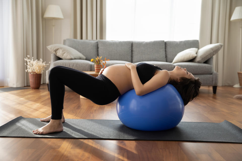 Concentrated pregnant woman doing exercises with fitness ball at home, practicing yoga, pilates with support on fitball, training body, core muscles. Sport activity in pregnancy, prenatal care concept