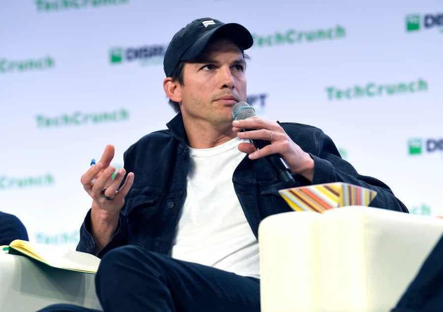 Ashton in a cap, sitting down, and holding a microphone