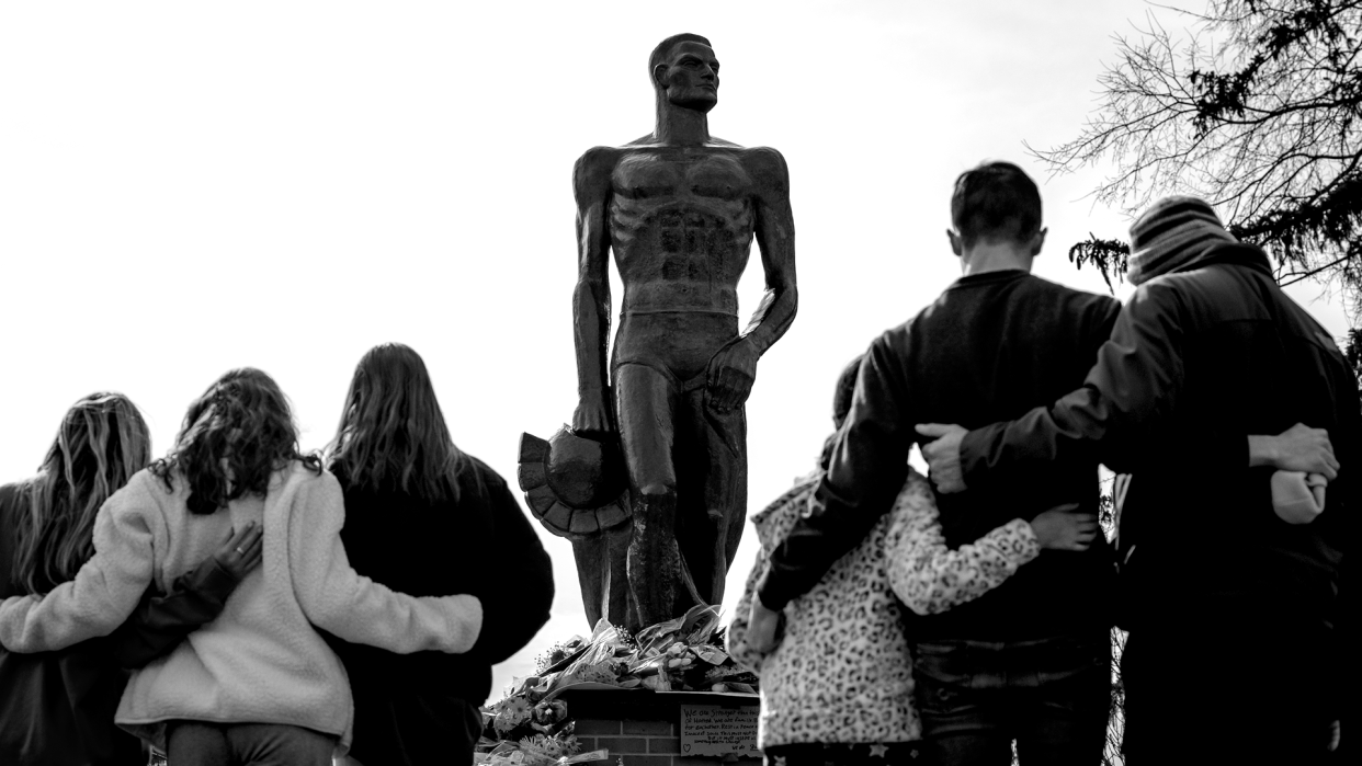 Seen with their backs to the camera against a bronze statue, students and families link arms and bow their heads.